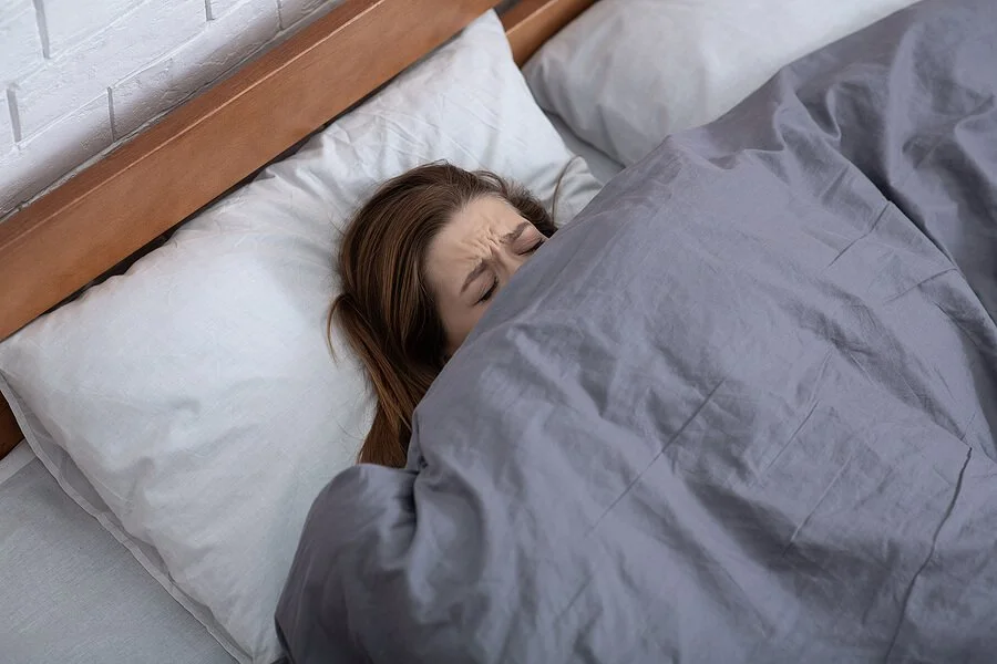 Woman laying in bed, looking stressed and upset. Symptoms of anxiety can feel overwhelming. Get anxiety treatment in Austin, TX from a skilled anxiety therapist via online therapy in Texas in Houston, Dallas, Austin, and beyond.