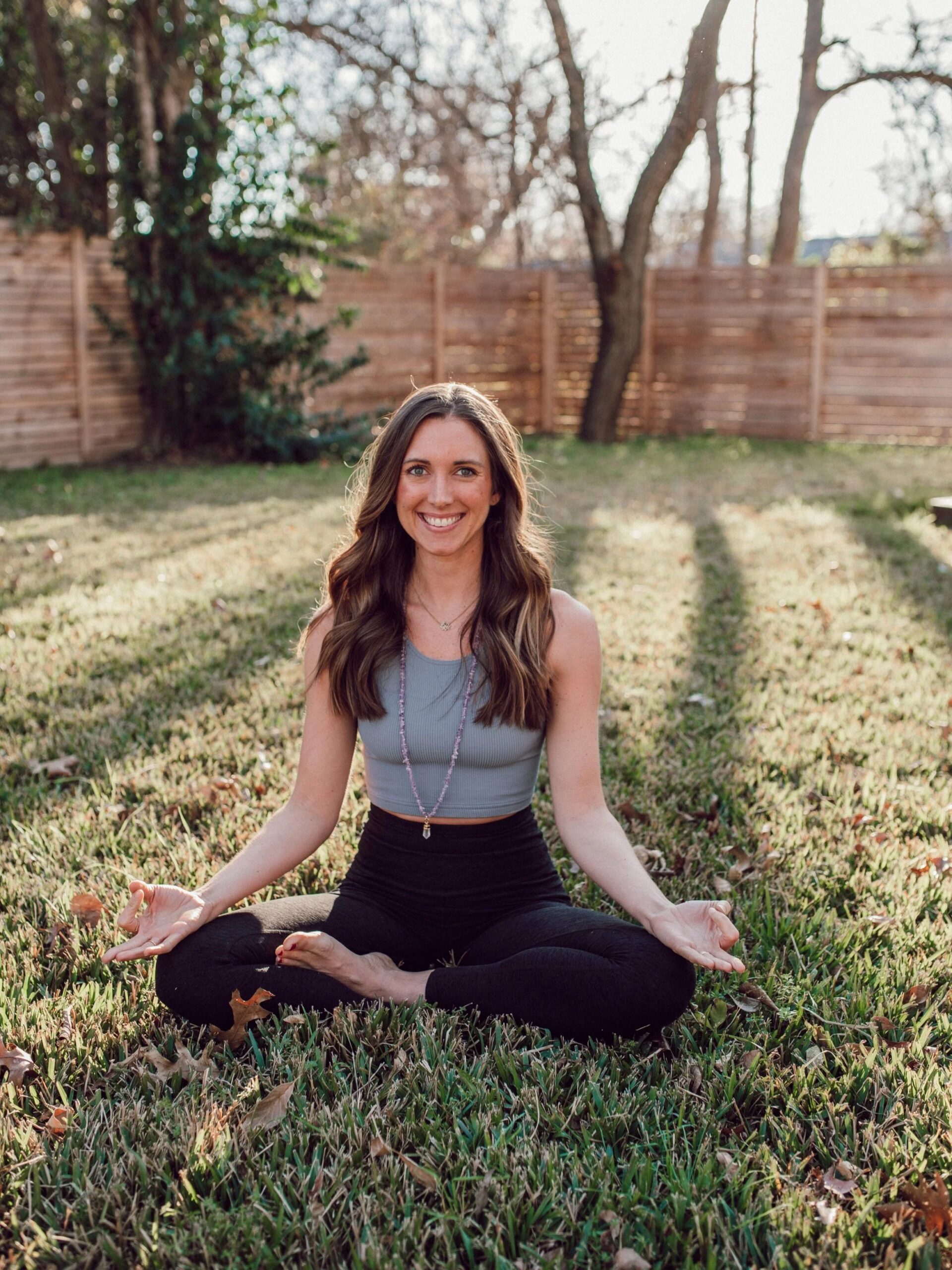 Image of Grace Mackey, a yoga therapist, sitting with her legs crossed and hands held in front of her while smiling at the camera. Working with a yoga therapist in Texas using therapeutic yoga can help you get in touch with yourself. Consider s…